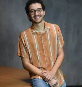 Foto de estúdio, à meia altura, de um jovem adulto, homem, de pele clara e cabelo escuro cacheado, com barba rala, usando óculos de aro fino e preto. Ele está sentado em uma mesa. O homem veste uma camisa casual de botões, de manga curta, na cor laranja com listras finas em um tom mais claro, e calça jeans azul-claro. Ele está sorrindo, olhando diretamente para a câmera, com as mãos cruzadas e apoiadas no colo. O fundo é uma parede cinza-escura, simples, com iluminação de estúdio.