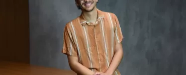 Foto de estúdio, à meia altura, de um jovem adulto, homem, de pele clara e cabelo escuro cacheado, com barba rala, usando óculos de aro fino e preto. Ele está sentado em uma mesa. O homem veste uma camisa casual de botões, de manga curta, na cor laranja com listras finas em um tom mais claro, e calça jeans azul-claro. Ele está sorrindo, olhando diretamente para a câmera, com as mãos cruzadas e apoiadas no colo. O fundo é uma parede cinza-escura, simples, com iluminação de estúdio.