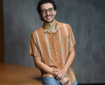 Foto de estúdio, à meia altura, de um jovem adulto, homem, de pele clara e cabelo escuro cacheado, com barba rala, usando óculos de aro fino e preto. Ele está sentado em uma mesa. O homem veste uma camisa casual de botões, de manga curta, na cor laranja com listras finas em um tom mais claro, e calça jeans azul-claro. Ele está sorrindo, olhando diretamente para a câmera, com as mãos cruzadas e apoiadas no colo. O fundo é uma parede cinza-escura, simples, com iluminação de estúdio.