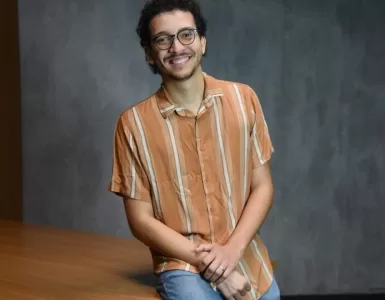 Foto de estúdio, à meia altura, de um jovem adulto, homem, de pele clara e cabelo escuro cacheado, com barba rala, usando óculos de aro fino e preto. Ele está sentado em uma mesa. O homem veste uma camisa casual de botões, de manga curta, na cor laranja com listras finas em um tom mais claro, e calça jeans azul-claro. Ele está sorrindo, olhando diretamente para a câmera, com as mãos cruzadas e apoiadas no colo. O fundo é uma parede cinza-escura, simples, com iluminação de estúdio.