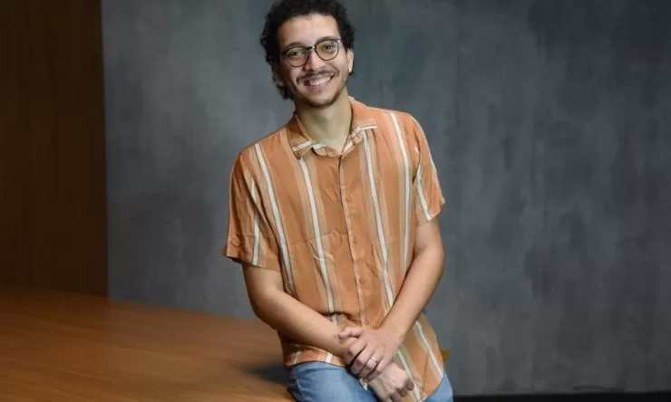 Foto de estúdio, à meia altura, de um jovem adulto, homem, de pele clara e cabelo escuro cacheado, com barba rala, usando óculos de aro fino e preto. Ele está sentado em uma mesa. O homem veste uma camisa casual de botões, de manga curta, na cor laranja com listras finas em um tom mais claro, e calça jeans azul-claro. Ele está sorrindo, olhando diretamente para a câmera, com as mãos cruzadas e apoiadas no colo. O fundo é uma parede cinza-escura, simples, com iluminação de estúdio.
