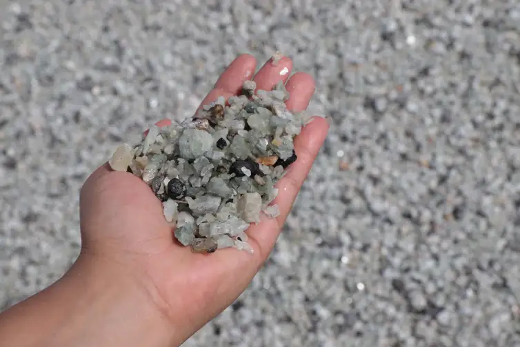 Uma fotografia capta uma mão de pele clara estendida, segurando um punhado de fragmentos de minério de lítio (provavelmente espodumênio). O material consiste em pedras pequenas, angulares e de cor predominante verde-claro a cinza-azulado pálido, algumas delas semitransparentes e misturadas com alguns pedaços escuros e opacos. O fundo da imagem está desfocado, mas coberto pelo mesmo material mineral, indicando que a foto foi tirada em uma pilha, no chão de uma mina ou em uma área de processamento.