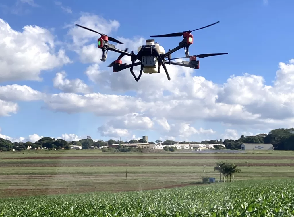 Um drone branco de grande porte com hastes pretas e detalhes em vermelho sobrevoando uma plantação enquanto aplica defensivos agrícolas. É dia com o céu azul e muitas nuvens espalhadas, ao fundo é possível ver galpões brancos e árvores.