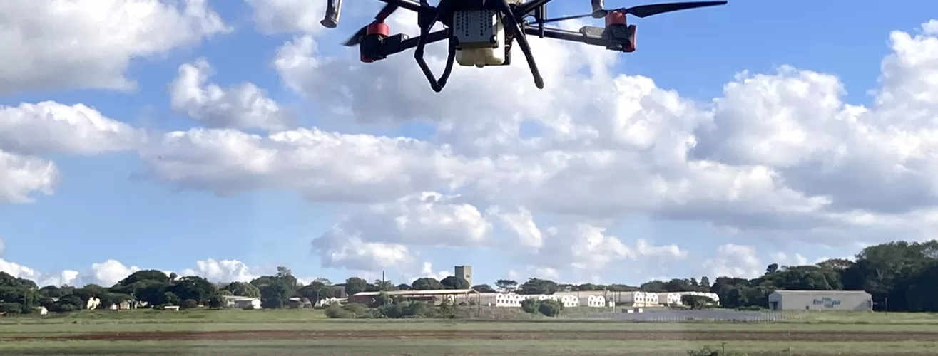 Um drone branco de grande porte com hastes pretas e detalhes em vermelho sobrevoando uma plantação enquanto aplica defensivos agrícolas. É dia com o céu azul e muitas nuvens espalhadas, ao fundo é possível ver galpões brancos e árvores.