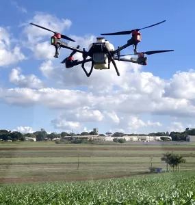 Um drone branco de grande porte com hastes pretas e detalhes em vermelho sobrevoando uma plantação enquanto aplica defensivos agrícolas. É dia com o céu azul e muitas nuvens espalhadas, ao fundo é possível ver galpões brancos e árvores.