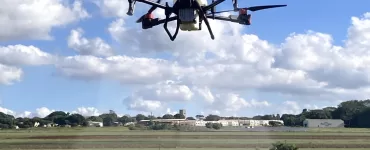Um drone branco de grande porte com hastes pretas e detalhes em vermelho sobrevoando uma plantação enquanto aplica defensivos agrícolas. É dia com o céu azul e muitas nuvens espalhadas, ao fundo é possível ver galpões brancos e árvores.