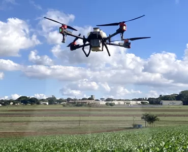 Um drone branco de grande porte com hastes pretas e detalhes em vermelho sobrevoando uma plantação enquanto aplica defensivos agrícolas. É dia com o céu azul e muitas nuvens espalhadas, ao fundo é possível ver galpões brancos e árvores.