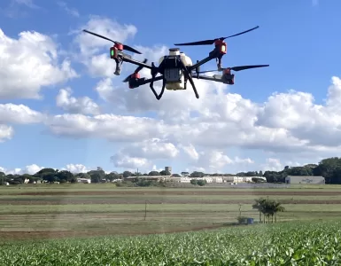 Um drone branco de grande porte com hastes pretas e detalhes em vermelho sobrevoando uma plantação enquanto aplica defensivos agrícolas. É dia com o céu azul e muitas nuvens espalhadas, ao fundo é possível ver galpões brancos e árvores.