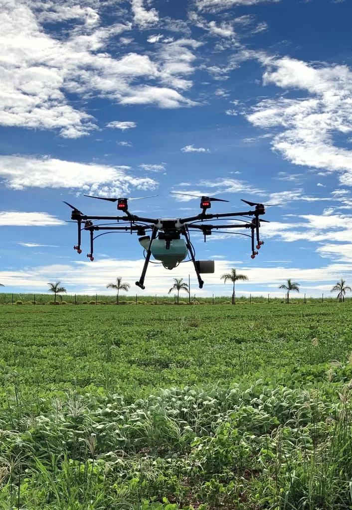 Um drone de grande porte sobrevoando uma plantação. É dia com o céu azul e as nuvens estão espalhadas, todo o chão é coberto por plantações até o horizonte e é possível ver algumas palmeiras enfileiradas ao fundo.
