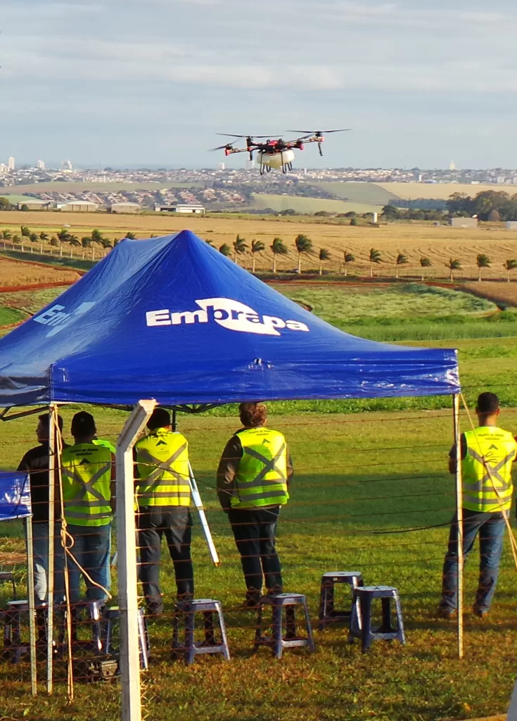 Um drone branco com hastes pretas faz um voo de demonstração em frente à uma tenda da Embrapa. A tenda tem um tecido azul com o logo da Embrapa em branco. Dentro dela há cinco homens vestindo coletes verdes para identificação. Eles estão sob campos verdes e, ao fundo, é possível ver áreas de plantio e palmeiras enfileiradas. Mais ao fundo é possível ver uma cidade.
