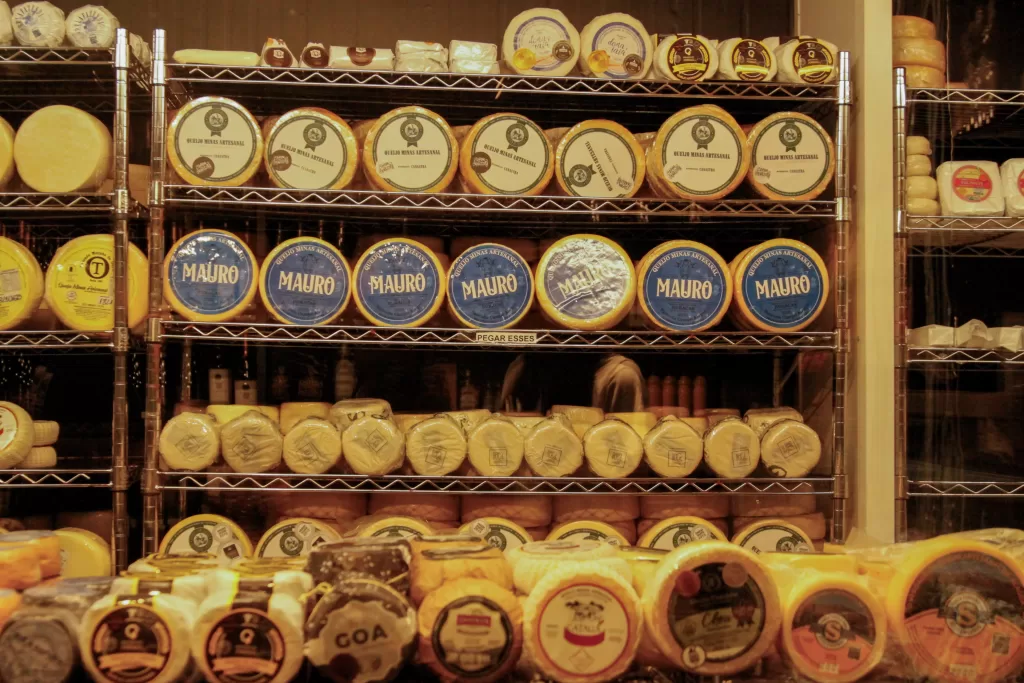 Prateleiras de metal cheias de queijos artesanais redondos e embalados, organizados em várias fileiras. As embalagens são principalmente em tons de amarelo, azul e branco, com rótulos visíveis como “Queijo Minas Artesanal” e “Mauro”. A vitrine é iluminada com luz quente, dando um tom amarelado à cena. Na parte inferior, há ainda mais queijos empilhados, criando a sensação de uma loja especializada em produtos artesanais.