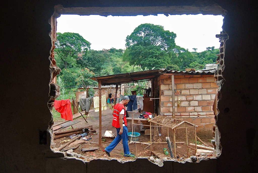 Um funcionário da prefeitura vestindo um colete vermelho vibrante avalia o estado de uma casa de tijolos. A casa é rodeada por árvores e mato. Essa cena é vista pelo buraco de uma janela.