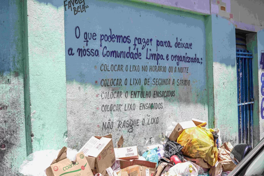 Um mural pintado em uma parede no Morro do Papagaio. O mural é verde, e abaixo dele estão várias sacolas de lixo e caixas de papelão. No mural, está escrito: "O que podemos fazer para deixar a nossa comunidade limpa e organizada: colocar o lixo no horário ou à noite; colocar o lixo de segunda a sábado; colocar o entulho ensacado; colocar lixos ensacados; não rasgue o lixo."