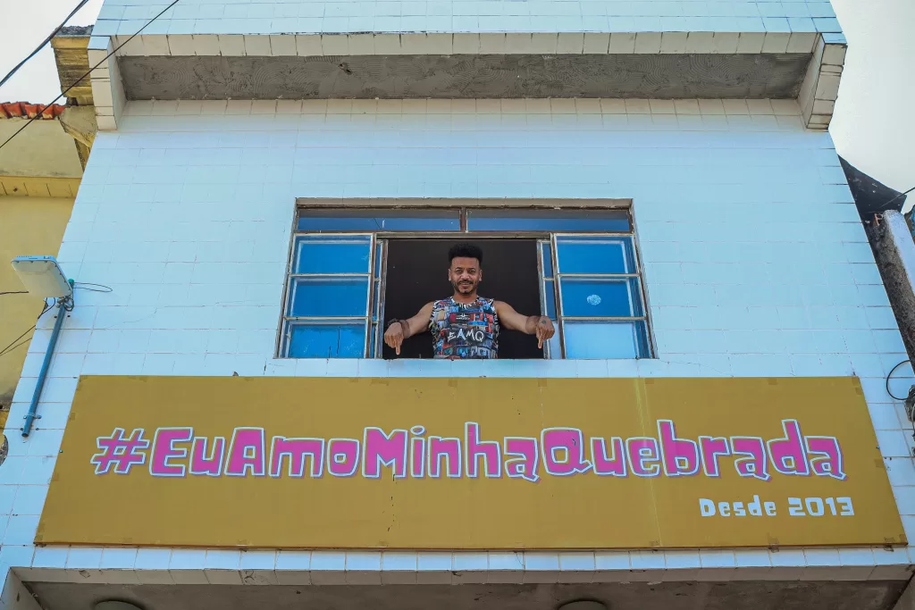 Júlio Fessô, um homem negro, aparece pela janela de um pequeno prédio branco. Ele aponta para um cartaz abaixo da janela, em que está escrito #EuAmoMinhaQuebrada