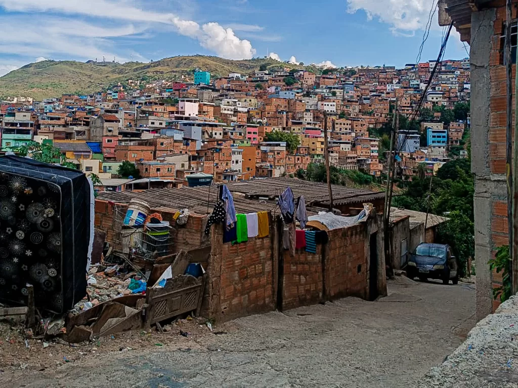 Uma rua asfaltada e suja de terra. Nela, avista-se um muro de tijolos, pertencente a uma pequena casa, lotado de roupas secando. Da rua, é possível observar a paisagem no Aglomerado da Serra: várias casas de tijolos, algumas pintadas. Elas estão empilhadas uma em cima da outra. Poucas árvores fazem parte da vista.