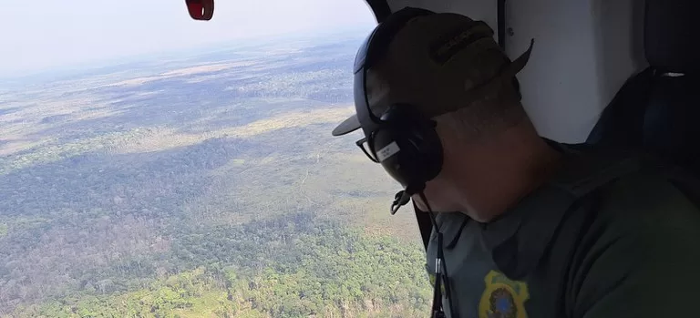 Agente do Ibama, visto de costas usando fones de ouvido e uniforme, observa de dentro de um helicóptero uma vasta paisagem de floresta intercalada com grandes áreas desmatadas.