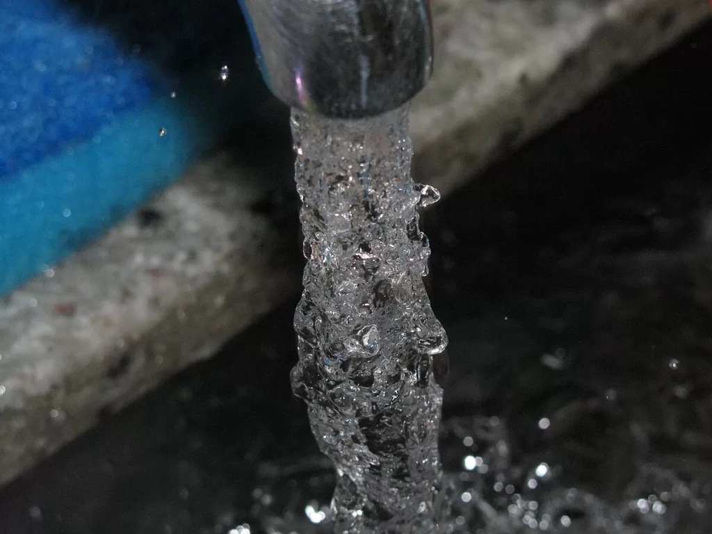 Um close-up de água fluindo de uma torneira metálica. A água é cheia de bolhas e parece uma massa cristalina e texturizada enquanto cai, criando respingos e ondas na superfície escura abaixo. O fundo desfocado mostra uma borda de granito cinza e uma área azul difusa à esquerda.