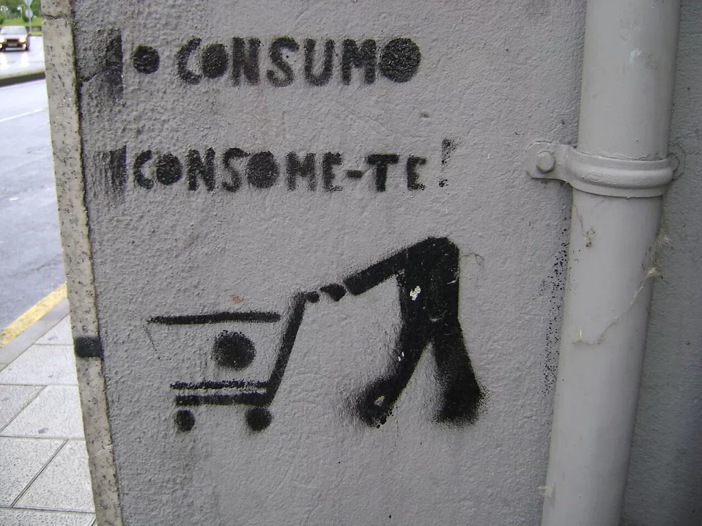 Uma foto de uma parede de concreto cinza com uma pichação em stencil preta. O grafite mostra o texto "O CONSUMO" na parte superior, seguido de "CONSOME-TE!" abaixo. Na parte inferior, há um desenho de uma figura humana empurrando um carrinho de compras, inclinada para frente como se estivesse sendo "consumida" pelo carrinho. À direita, um tubo de encanamento vertical cinza está fixado à parede. O fundo à esquerda mostra uma rua molhada e um carro branco passando em um dia nublado.