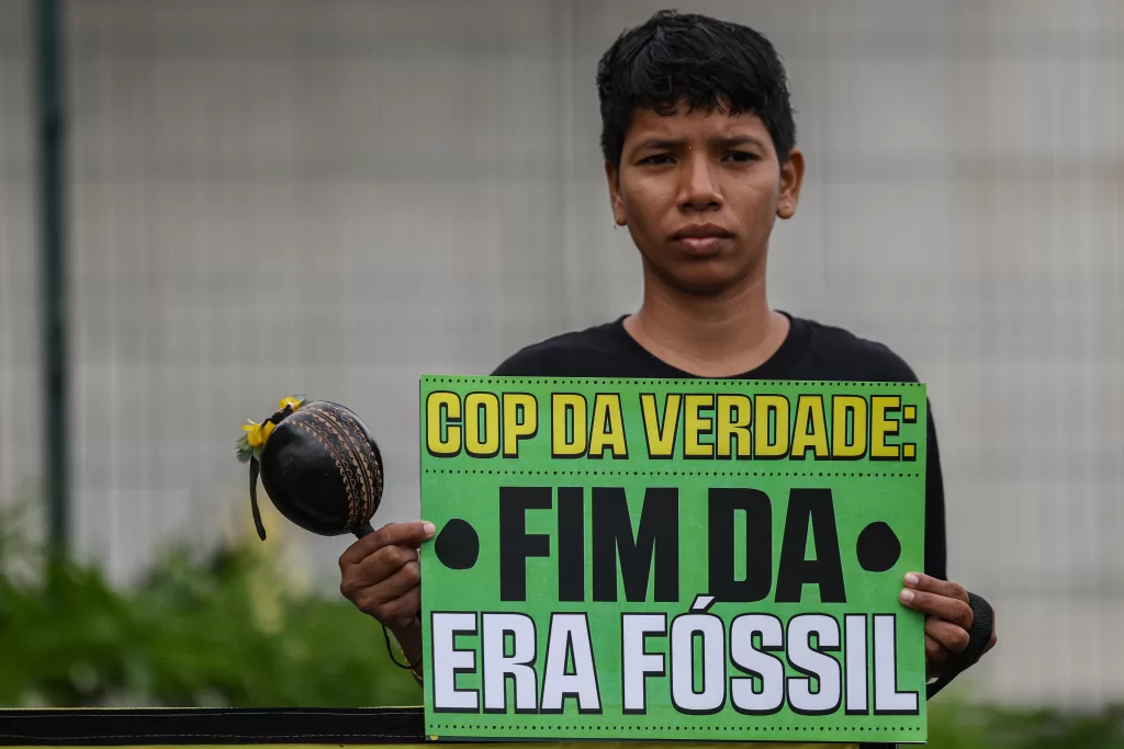 Fotografia em plano médio de um jovem indígena de cabelos curtos e escuros e expressão séria, vestindo uma camiseta preta. Ele segura na altura do peito um cartaz verde com a mensagem em letras garrafais pretas e brancas: 'COP DA VERDADE: FIM DA ERA FÓSSIL'. Em sua mão direita, ele segura também um maracá preto com detalhes amarelos. O jovem olha diretamente para a câmera. O fundo é cinza e desfocado, sugerindo uma grade ou fachada de prédio.