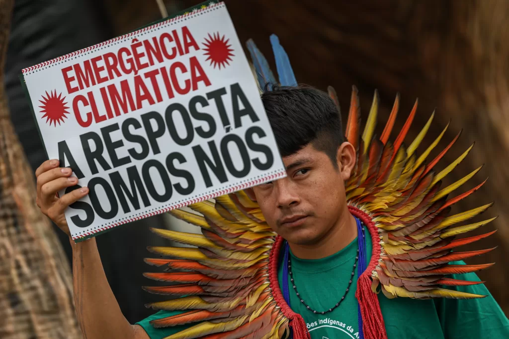 Indígena segurando um cartaz branco com letras pretas e vermelhas onde se lê: 'EMERGÊNCIA CLIMÁTICA A RESPOSTA SOMOS NÓS'. O cartaz possui detalhes gráficos vermelhos semelhantes a sóis nos cantos. O indígena usa um grande cocar de penas em tons de amarelo, laranja e vermelho que emoldura sua cabeça e ombros, além de um colar escuro e camiseta verde. Ele olha para a lateral esquerda, fora do enquadramento. O fundo está desfocado.