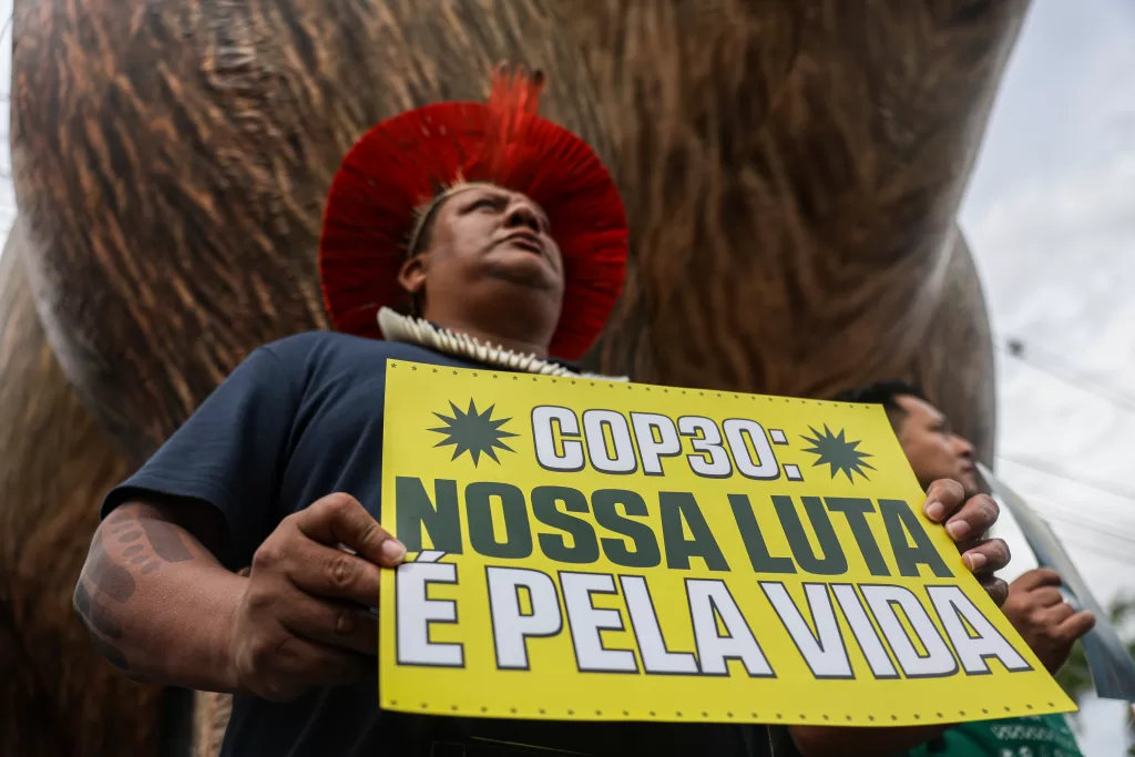 Foto em ângulo baixo de um homem indígena segurando, com as duas mãos, um cartaz amarelo com os dizeres em letras garrafais verdes e brancas: 'COP30: NOSSA LUTA É PELA VIDA'. O homem usa uma camiseta azul-escura, um colar grosso de contas brancas e um cocar de penas vermelhas. Ele olha para o alto e para a direita com expressão séria. Ao fundo, vê-se o céu nublado e, desfocada, parte de uma grande estrutura marrom.