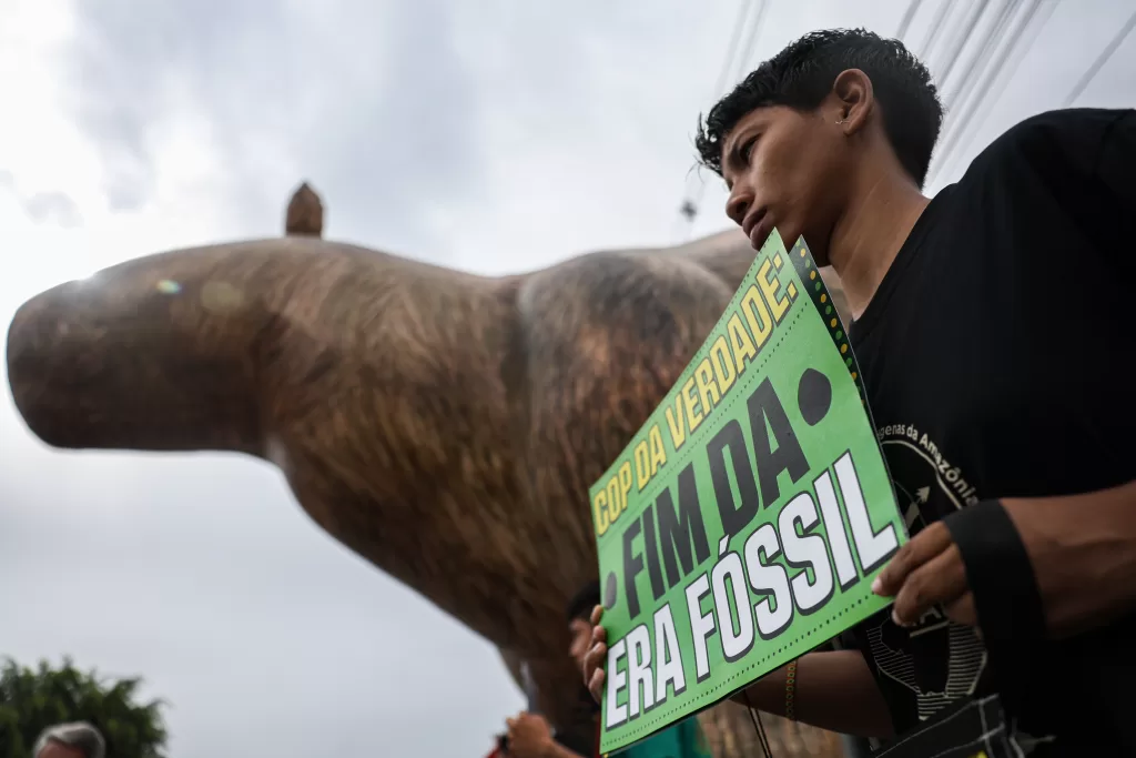 Foto em ângulo baixo de uma pessoa jovem de perfil, segurando um cartaz verde com letras garrafais que dizem: 'COP DA VERDADE: FIM DA ERA FÓSSIL'. Ao fundo, vê-se parcialmente um grande boneco inflável marrom (semelhante a uma capivara ou animal de grande porte) contra um céu nublado.