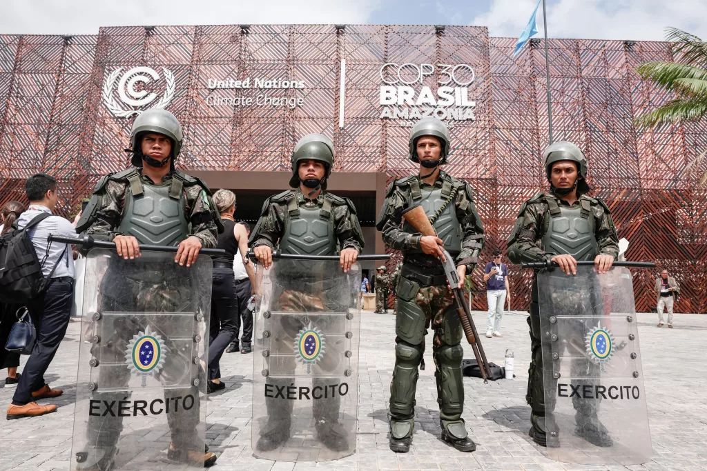Fotografia jornalística horizontal. Em primeiro plano, quatro soldados do Exército Brasileiro estão em pé, lado a lado, fazendo a segurança em frente a um edifício. Eles vestem uniformes camuflados em tons de verde, capacetes e coletes de proteção balística. Três deles apoiam no chão grandes escudos transparentes onde se vê o brasão da República e a palavra "EXÉRCITO" em letras maiúsculas; o terceiro soldado (da esquerda para a direita) empunha uma arma longa. Ao fundo, vê-se a fachada de uma estrutura coberta por treliças de madeira, com letreiros brancos que dizem "United Nations Climate Change" e "COP 30 BRASIL AMAZÔNIA". Algumas pessoas civis caminham atrás dos soldados.