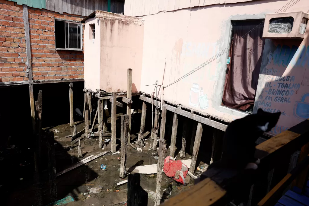 Fotografia colorida em plano médio que retrata construções de palafitas na Comunidade da Vila da Barca, em Belém. A imagem mostra o vão entre duas casas: à esquerda, uma parede de tijolos vermelhos aparentes com uma janela de alumínio gradeada; à direita, uma parede de alvenaria pintada de rosa claro, com uma porta coberta por uma cortina marrom e dizeres escritos à mão na parede em tinta azul, listando itens como "bolsa", "toalha" e "brincos".
As estruturas são sustentadas por dezenas de estacas de madeira, muitas delas com aparência envelhecida e irregular, fincadas em um solo de lama escura onde se nota algum lixo acumulado. A iluminação é natural, com sombras fortes indicando sol intenso. No canto inferior direito, em primeiro plano e fora de foco, vê-se a silhueta de um gato preto sentado sobre um parapeito de madeira, observando.