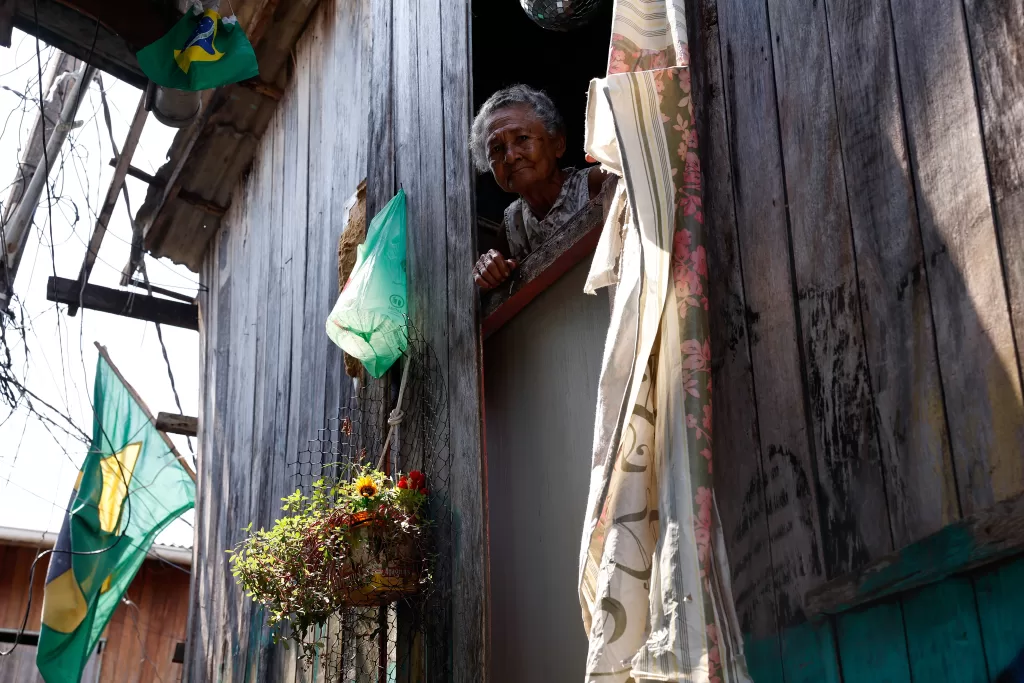 Fotografia vertical, em ângulo levemente baixo, de Cleonice da Silva Vera Cruz. Ela é uma senhora idosa de pele parda e cabelos grisalhos curtos, que aparece debruçada na abertura de uma janela em uma casa de madeira rústica. Cleonice olha diretamente para a câmera com uma expressão serena. A parede externa da casa é feita de tábuas verticais escuras e desgastadas. À esquerda da janela, há uma sacola plástica verde pendurada em um prego e, logo abaixo, um vaso suspenso com folhagens e flores amarelas e vermelhas. À direita, uma cortina clara com estampa floral rosa pende ao lado da abertura.