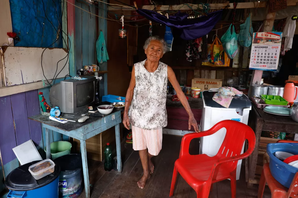 Fotografia de plano inteiro do interior da modesta casa de madeira de Cleonice da Silva Vera Cruz. Ela é uma mulher idosa, de cabelos grisalhos curtos e pele parda, que está de pé no centro do cômodo, olhando frontalmente para a câmera. Veste uma blusa branca sem mangas com estampa floral cinza e bermuda clara. Sua mão esquerda repousa sobre o encosto de uma cadeira de plástico vermelha vibrante. O ambiente está repleto de pertences: à esquerda, uma mesa de madeira azul sustenta uma televisão de tubo antiga, um relógio, remédios e um prato com comida; à direita, há uma máquina de lavar branca. O fundo está cheio de roupas e sacolas penduradas em varais, caixas de papelão e diversos utensílios domésticos.