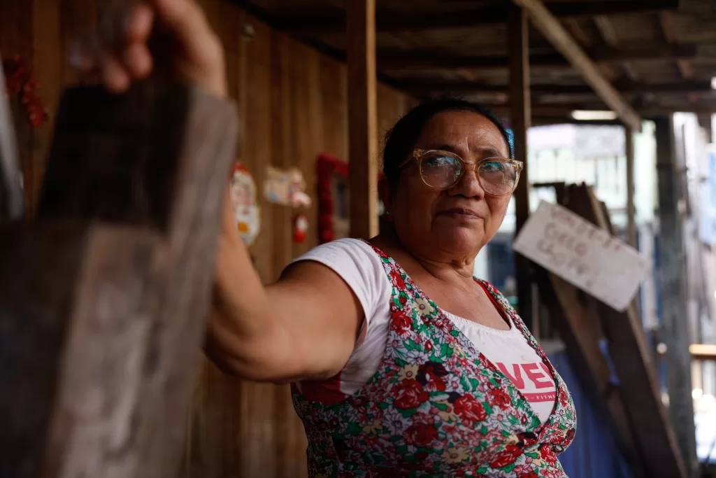 Fotografia em plano médio de Ivone Vidal, uma mulher de pele parda e cabelos escuros presos para trás. Ela usa óculos de grau com armação transparente e uma blusa branca com estampa floral vermelha. Ivone olha para a lateral direita com uma expressão serena e apoia o braço direito em uma viga de madeira rústica, que ocupa a lateral esquerda da foto. O fundo está desfocado, sugerindo o interior de uma casa de madeira com objetos pendurados.