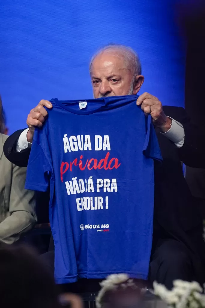 Presidente Lula segurando uma camisa azul escura escrito "Água da privada não dá para engolir", do Sindágua-MG, Sindicato dos Trabalhadores nas Indústrias de Purificação e Distribuição de Água e em Serviços de Esgotos de MG. A foto foi tirada em evento que contou com a presença do presidente em Belo Horizonte.