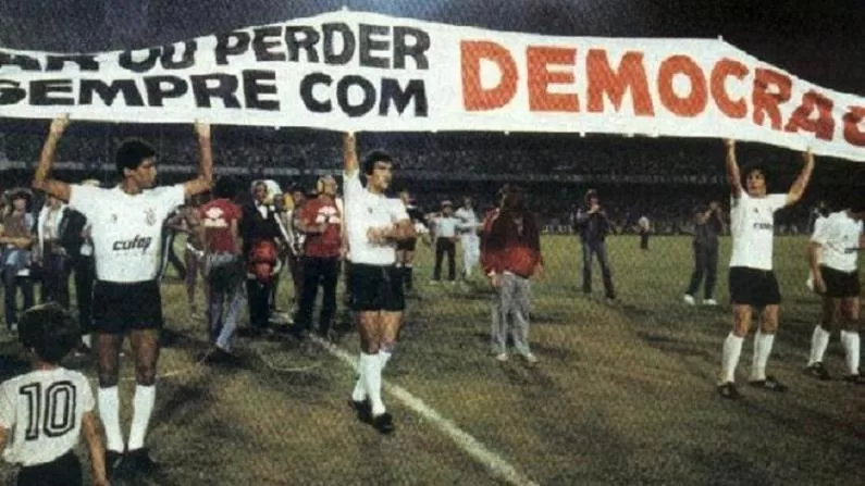Jogadores do Corinthians caminhando no gramado segurando uma grande faixa escrito: “Ganhar ou perder, mas sempre com Democracia”. Ao fundo, alguns fotógrafos capturam o momento.