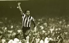 Jogador Reinaldo comemorando um gol com seu tradicional gesto de erguer um punho ao alto, torcida comemora ao fundo