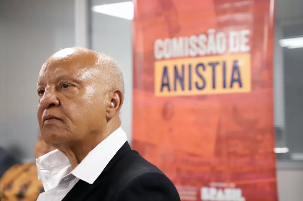 Reinaldo com expressão séria de lado e ao fundo um banner escrito “Comissão de Anistia”