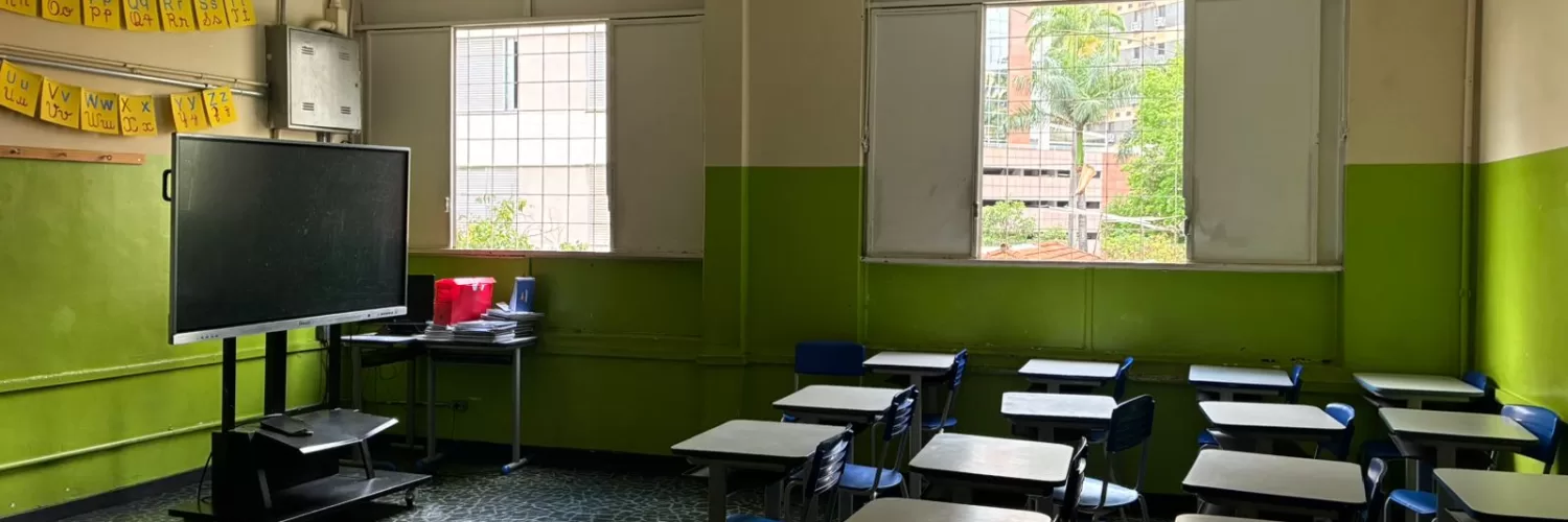 Uma sala de aula com carteiras azuis e brancas enfileiradas, duas janelas quadradas aberta deixando a luz entrar e um quadro negro móvel.