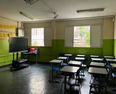 Uma sala de aula com carteiras azuis e brancas enfileiradas, duas janelas quadradas aberta deixando a luz entrar e um quadro negro móvel.