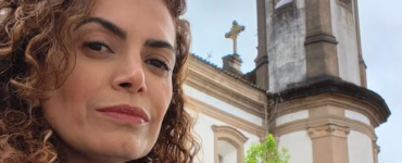 Na imagem tem uma mulher de cabelos escuros posando de frente, usando regata azul e um lenço branco e azul. Ao fundo, temos uma pequena árvore atrás da mulher e também igreja barroca histórica, pintada de amarelo nas bordas e branco como cor principal.