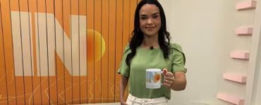 Usando uma blusa verde-claro e uma calça jeans bege-claro, a jornalista segura uma caneca branca com um sol e a letra “N” na frente.