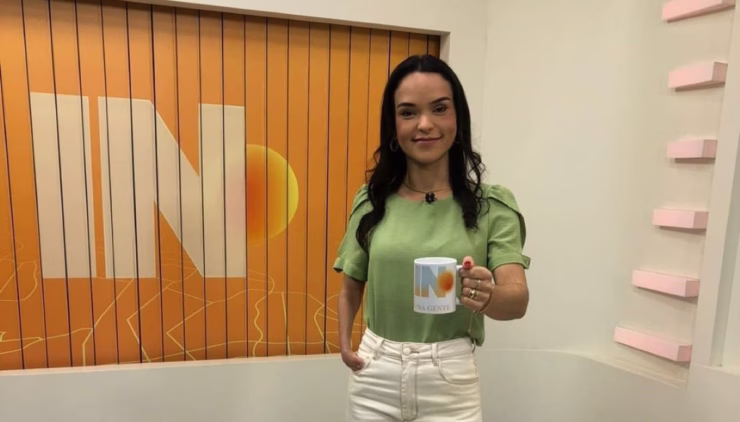 Usando uma blusa verde-claro e uma calça jeans bege-claro, a jornalista segura uma caneca branca com um sol e a letra “N” na frente.