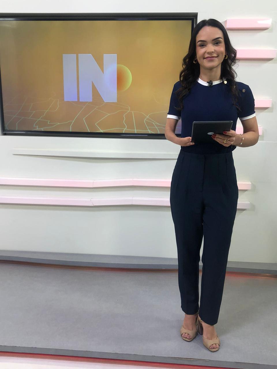 A jornalista usa um conjunto de calça e blusa azul-marinho e está em um estúdio de telejornal enquanto segura um tablet preto.
