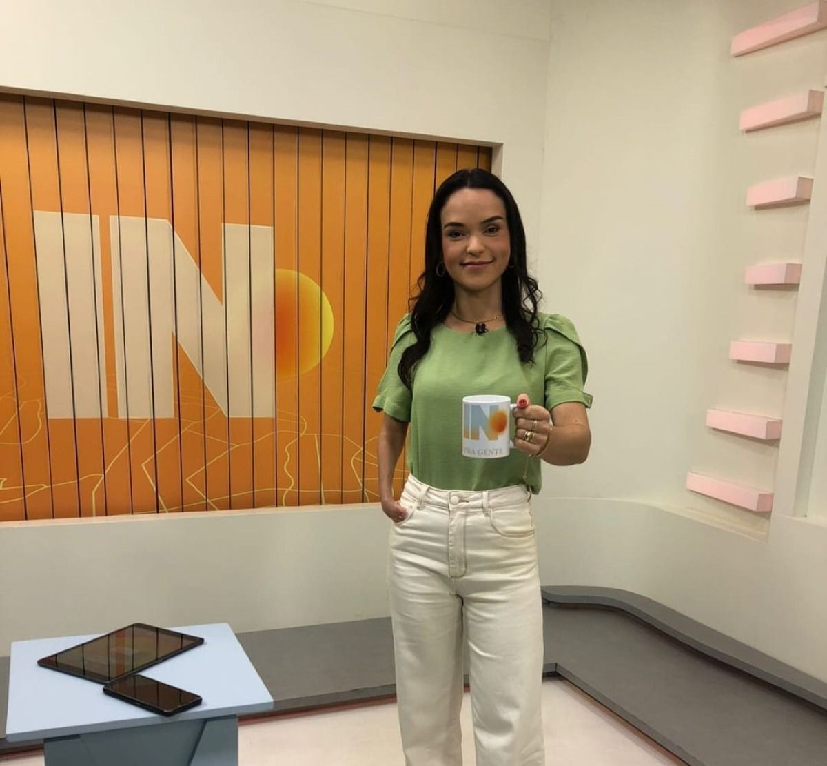 Usando uma blusa verde-claro e uma calça jeans bege-claro, a jornalista segura uma caneca branca com um sol e a letra “N” na frente.