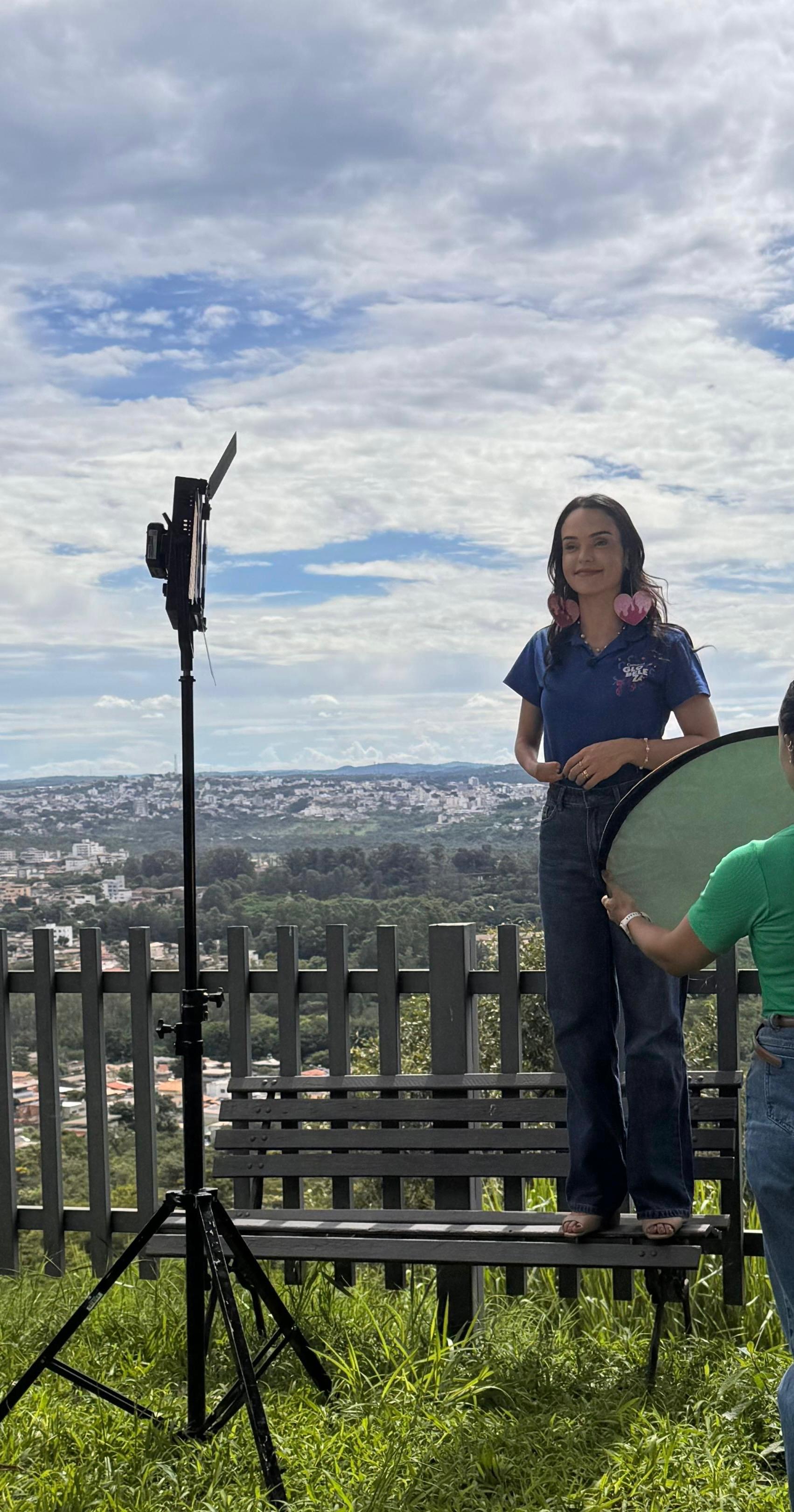 De pé em cima de um banco, a jornalista usa um conjunto de calça preta e camisa azul-escuro enquanto é cercada por equipamentos para gravação ao ar livre