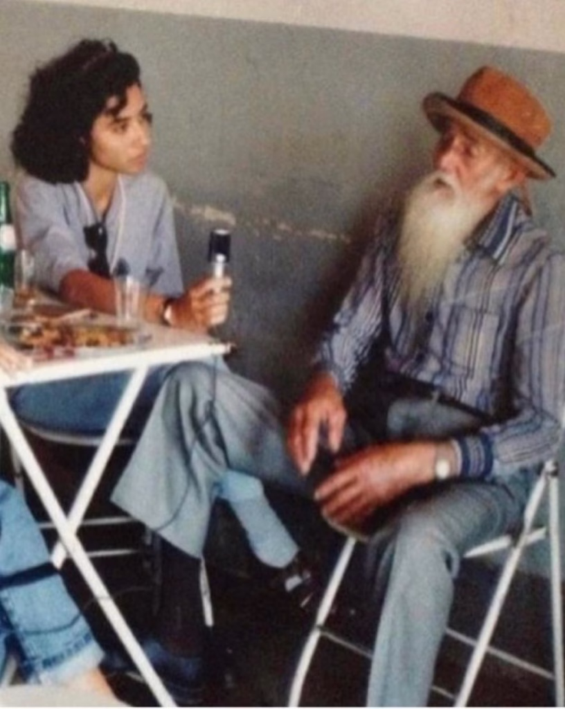 A foto retrata duas pessoas sentadas em cadeiras, com uma mesa de centro entre elas. À esquerda, Patrícia Pinheiro, com cabelo escuro e cacheado está sentada, vestindo uma camisa azul clara e calças cinzas. Ela segura um microfone na mão direita. À direita, um homem idoso com uma longa barba branca e um chapéu marrom está sentado. Ele veste uma camisa xadrez em tons de azul e branco e calças cinzas. Seus olhos estão fechados e sua expressão é serena. O fundo é uma parede de cor neutra.
