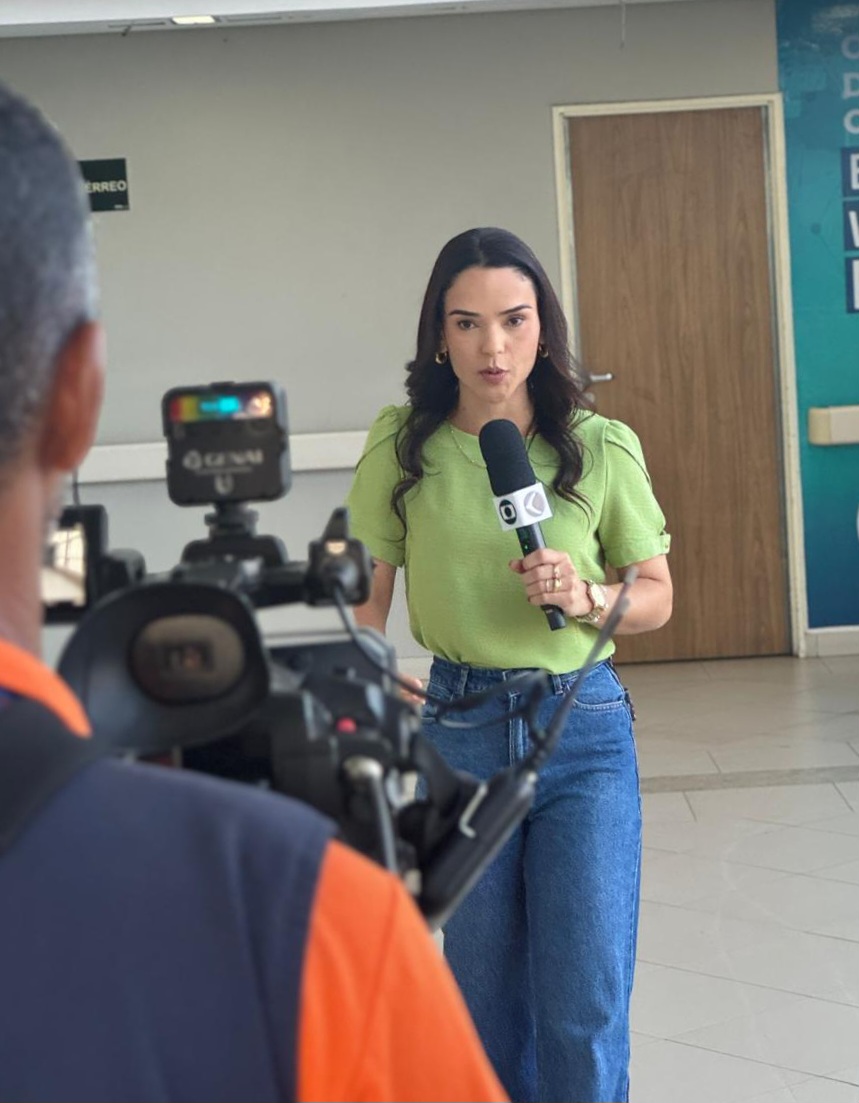 Vestindo uma blusa verde-claro e calça jeans, a apresentadora está de frente para a câmera enquanto conduz a reportagem.