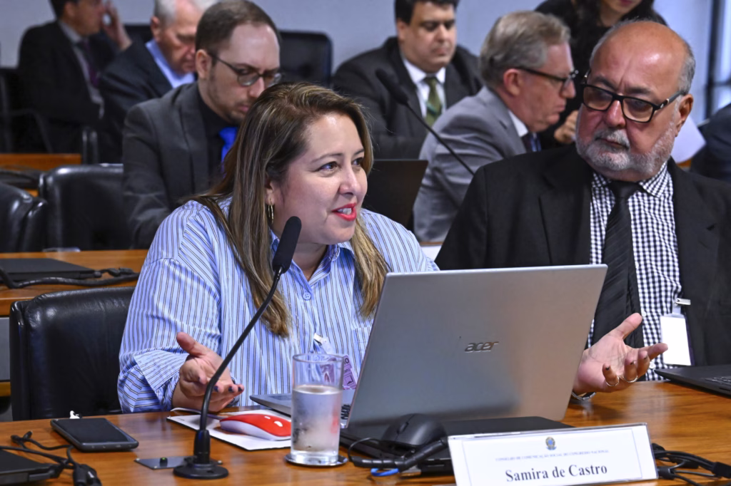 Samira de Castro, presidente da Federação Nacional dos Jornalistas (Fenaj), está sentada à mesa diante de um computador portátil e de um microfone. Ela usa uma camisa azul listrada e batom vermelho, enquanto fala e gesticula com as mãos.
