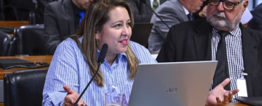 Samira de Castro, presidente da Federação Nacional dos Jornalistas (Fenaj), está sentada à mesa diante de um computador portátil e de um microfone. Ela usa uma camisa azul listrada e batom vermelho, enquanto fala e gesticula com as mãos.