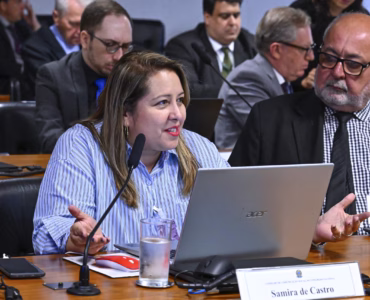 Samira de Castro, presidente da Federação Nacional dos Jornalistas (Fenaj), está sentada à mesa diante de um computador portátil e de um microfone. Ela usa uma camisa azul listrada e batom vermelho, enquanto fala e gesticula com as mãos.