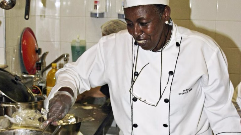Chef mulher negra sorrindo enquanto mexe alimento em panela dentro de cozinha profissional