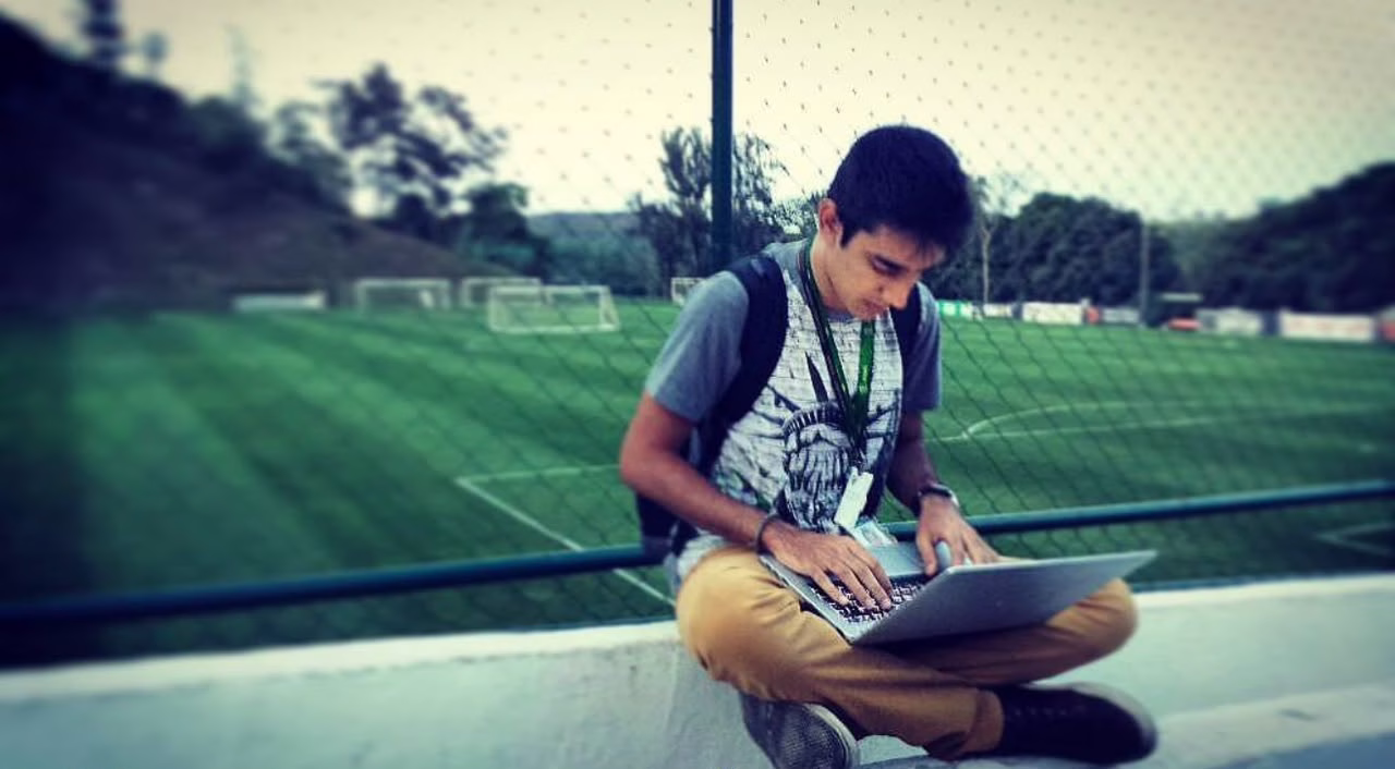 Sentado na arquibancada com um campo de futebol ao fundo, Guilherme está escrevendo em um notebook no colo.